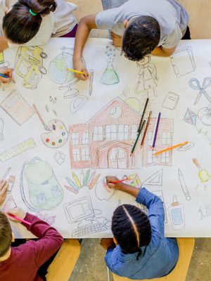 An aerial view of a multi ethnic group of children colour in a series of back to school related images. The artwork includes pictures of a backpack, microscope, paints, bus, scissors, art supplies, and books.